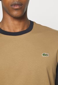 Gros plan d'une personne portant une chemise beige avec un col noir et un petit logo crocodile vert sur la poitrine.
