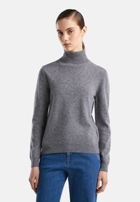 Donna che indossa un maglione grigio a collo alto e jeans blu, in piedi contro uno sfondo chiaro e uniforme, guardando leggermente di lato.