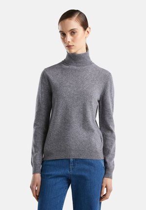 Femme portant un pull à col roulé gris et un jean bleu, se tenant devant un fond uni clair, regardant légèrement sur le côté.