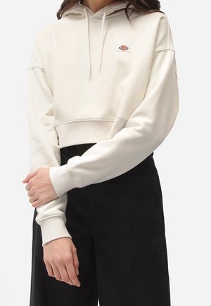 Personne portant un sweat à capuche court crème avec cordons et un petit logo sur la poitrine, associé à un pantalon noir taille haute, bras croisés.