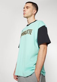 Camiseta verde menta claro con mangas cortas negras, que presenta la palabra "CHARLOTTE" en letras doradas en el pecho. Textura de tela suave.