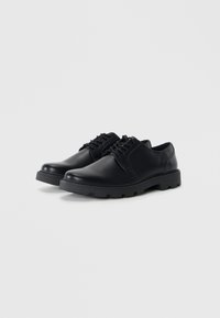 Zapatos de cuero negro con cordones, punta redondeada, suela texturizada y parte superior lisa. Presentan un minimalismo en las costuras y un diseño robusto.