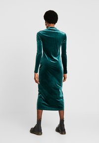 Vestido midi de terciopelo en color verde azulado con mangas largas y cuello alto, que presenta una silueta ajustada y un brillo sutil. Combinado con botas negras de tobillo.