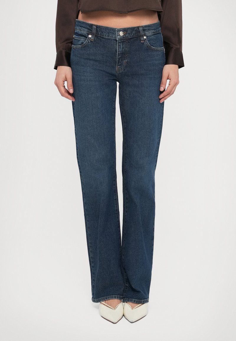 Elölnézet egy személyről, aki sötétkék, egyenes szárú farmert, barna rövid felsőt és fehér hegyes orrú cipőt visel, fehér háttér előtt.