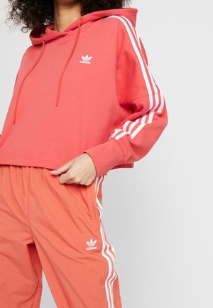 Osoba w koralowej, krótkiej bluzie z kapturem Adidas i pasujących spodniach dresowych z białymi paskami i logo, ręka spoczywa blisko kieszeni.