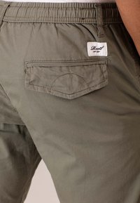 Pantalones verde oliva con una cintura elástica, un bolsillo trasero con tapa y una etiqueta de marca visible cosida en el bolsillo.