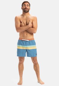 Bañadores de hombre en azul con rayas horizontales amarillas y verdes, con cinturilla elástica y cordón, y pequeño logo en la pierna.