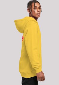 Yellow hooded sweatshirt with long sleeves. Fabric appears soft, featuring a front pocket and a graphic design in red on the left side.