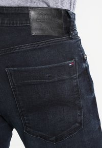 Mörka denimjeans med en svart läderetikett som säger "TOMMY JEANS." Funktioner inkluderar en bakficka med diskret sömnad och en liten flaggaccent.
