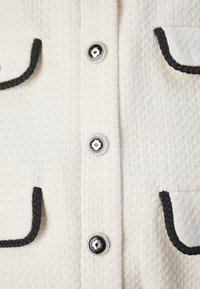 Gros plan sur un tissu texturé crème avec un galon tressé noir sur les poches et des boutons décoratifs noirs et argentés.