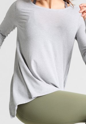Hellgraues Langarmoberteil in lockerer Passform mit Rundhalsausschnitt und seitlichen Schlitzen, kombiniert mit olivgrünen Leggings. Das Material wirkt weich.