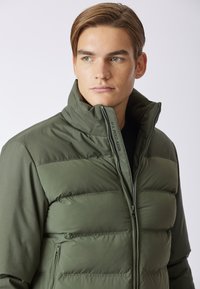 Giacca puffer verde oliva con alto colletto, chiusura a zip e dettaglio del logo sulla zip. Caratterizzata da sezioni trapuntate per l'isolamento.