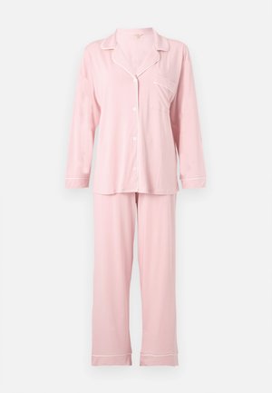 Mujer de pie descalza, vistiendo un pijama rosa claro de manga larga con botones y pantalones anchos a juego sobre fondo blanco.