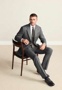 Grauer Wollanzug mit Kerbkragen, weißem Hemd und gemustertem Krawatte. Schwarze Lederschuhe, sitzend auf einem Holzstuhl vor neutralem Hintergrund.