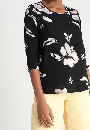 Chemise noire avec imprimé floral rose pâle et blanc, manches trois quarts, portée avec un pantalon jaune clair et une bague en or à la main gauche.