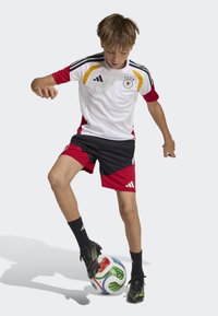 Adidas youth soccer uniform with a white top featuring red and yellow accents, black shorts with red and black design, and black socks, playing with a multicolored ball.