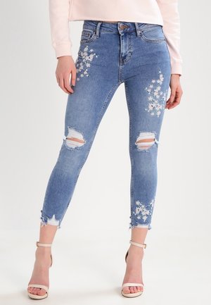 Jean skinny bleu avec genoux déchirés et broderie florale blanche, porté avec des sandales à talons beiges et un top rose pâle.