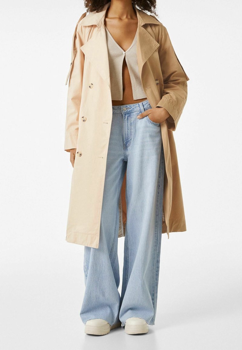 Beżowy trencz z szerokim kołnierzem i guzikami, w połączeniu z jasnoszarym topem typu crop i jasnoniebieskimi spodniami dzwonami, noszony z białymi butami.