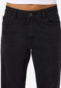 Detaj blerjët zgjidhje e belit të mesëm e veshur me xhinse të zeza me bel të lartë, me mbyllje me dügë dhe tërheqës, të kombinuara me një bluzë të bardhë të shkurtër.