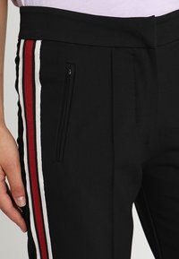 Svarta joggers med sidofickor, med röda, vita och marinblå ränder längs yttersemmarna. Tillverkade av ett mjukt, elastiskt material.