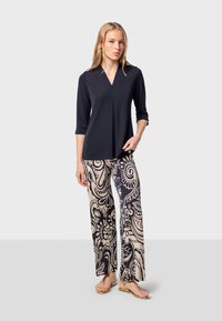 Marinblus med trekvartsärmar, V-ringning och veckdetalj, ihop med beiga vida byxor med en mörk blommig swirl-mönster. Guldtofflor.