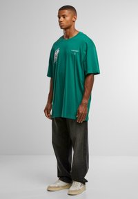 Green oversized cotton t-shirt with graphic print on the chest, paired with dark jeans and light-colored sneakers. Neutral background.