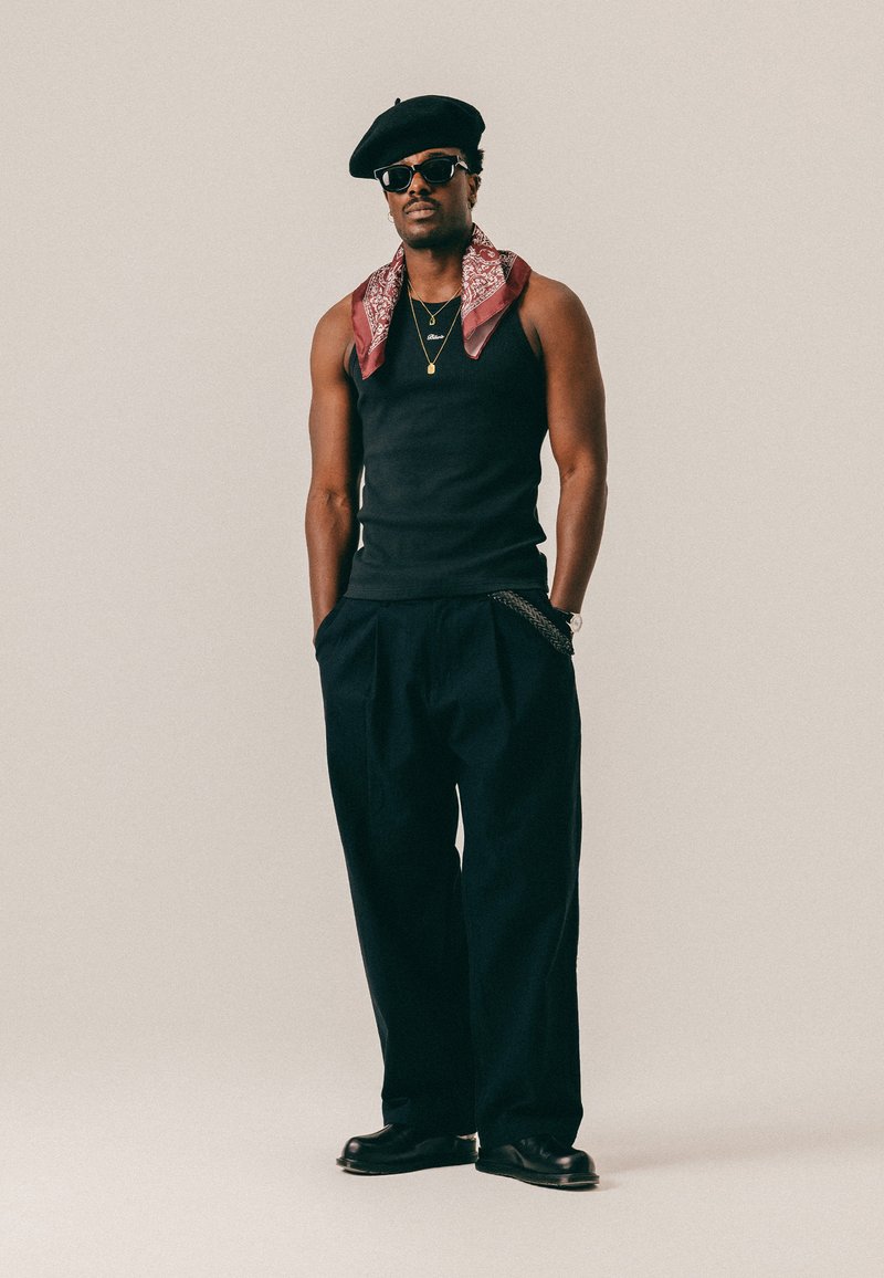 Homme portant un béret noir, des lunettes de soleil, un débardeur noir, un pantalon large noir, un bandana rouge autour du cou et des chaussures noires, debout les mains dans les poches.