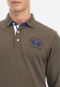 Barna pólóing kék gallérral, fehér csíkokkal a nyakrészen és kék hímzett logóval, amely lovasokat és szöveget ábrázol.