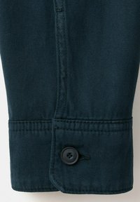 Poignet de manche en denim bleu foncé avec double couture et un seul bouton noir fixé à travers un boutonnière.