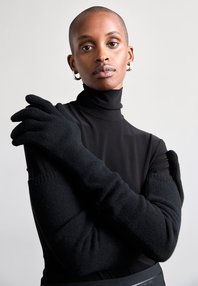 Sorte strikkede handsker har et langt design med ribbede detaljer og en glat tekstur. Modellen bærer en tætsiddende sort højhalset bluse.