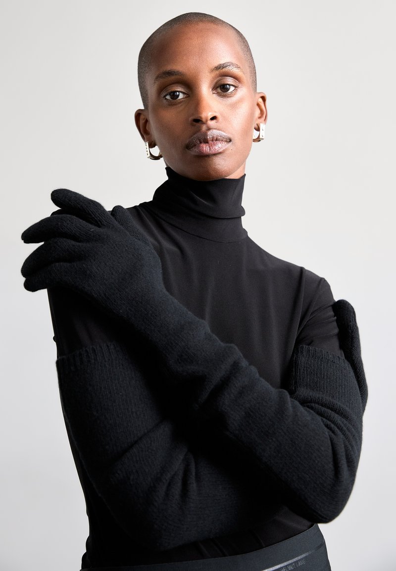 Mânușile negre tricotate au un design lung, cu accente ribbate și o textură netedă. Modelul poartă un top negru, cu guler înalt, ajustat.