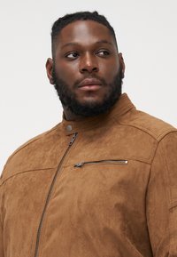 Veste en daim marron avec un col haut, fermeture éclair et une petite poche zippée sur la poitrine. Texture douce avec une finition lisse et un design minimaliste.