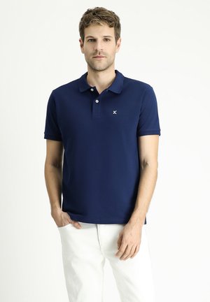 Marinefarbener Poloshirt aus glattem Stoff, mit Kragen, drei Knöpfen und einem kleinen Logo auf der linken Brust. Getragen mit weißen Hosen.