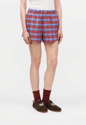 Person iført røde og blå ternede shorts, hvid ærmeløs top, mørkerøde sokker og brune ruskindssneakers står på en ensfarvet baggrund.