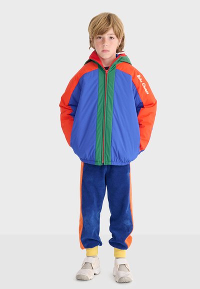 Niño con chaqueta con capucha azul, naranja y verde con el texto "Bobo Choses", pantalones azules y naranjas, calcetines amarillos y zapatillas blancas.