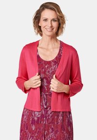 Fuchsia cardigan met driekwartmouwen, open voorkant en een gladde textuur, gedragen over een gestreepte paarse bloemenjurk.