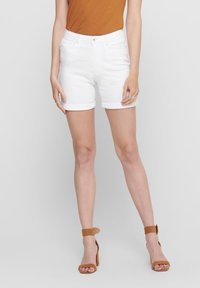 Shorts en denim blanc avec des ourlets retroussés, taille haute, et style à cinq poches. Associés à un haut marron à manches courtes et des sandales à talons hauts beiges.