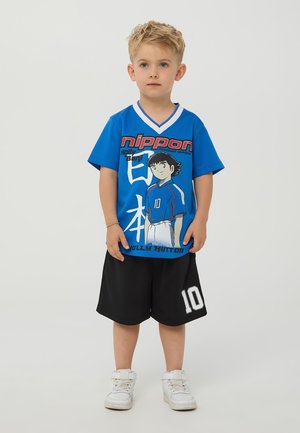 T-shirt blu in rete con stampa grafica di un personaggio, pantaloni corti neri con il numero 10 bianco e scarpe da ginnastica bianche con cinturini in velcro.