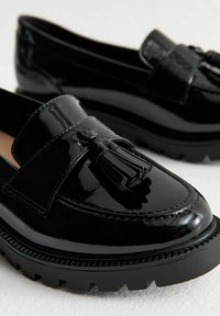 Mocassins en cuir verni noir avec un détail de pompon, dotés d'une semelle noire épaisse et de coutures le long du bord pour plus de texture et de détails.