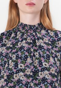 Blusa floreale con sfondo blu navy; presenta un design plissettato, un collo alto arricciato e una combinazione di motivi floreali rosa, bianchi e verdi.