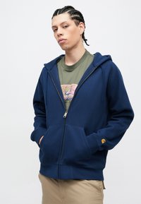 Tamsiai mėlynas džemperis su užtrauktuku, pagamintas iš minkštos medžiagos, su kišene priekyje ir briaunotomis rankogaliais. Dėvimas ant žalios grafinės marškinėlių.