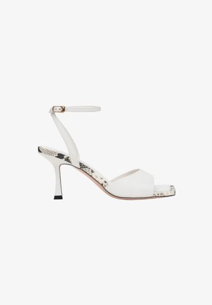 Bílé sandály na podpatku s otevřenou špičkou, řemínkem kolem kotníku a texturovaným detailem na podpatku s hadím vzorem. Podešev je světle zbarvená a design je minimalistický.