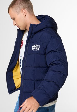 Junger Mann trägt eine marineblaue Red Bull Racing Steppjacke mit Kapuze, geöffnet, sodass das gelbe Jackenfutter und ein weißes Hemd sichtbar sind.