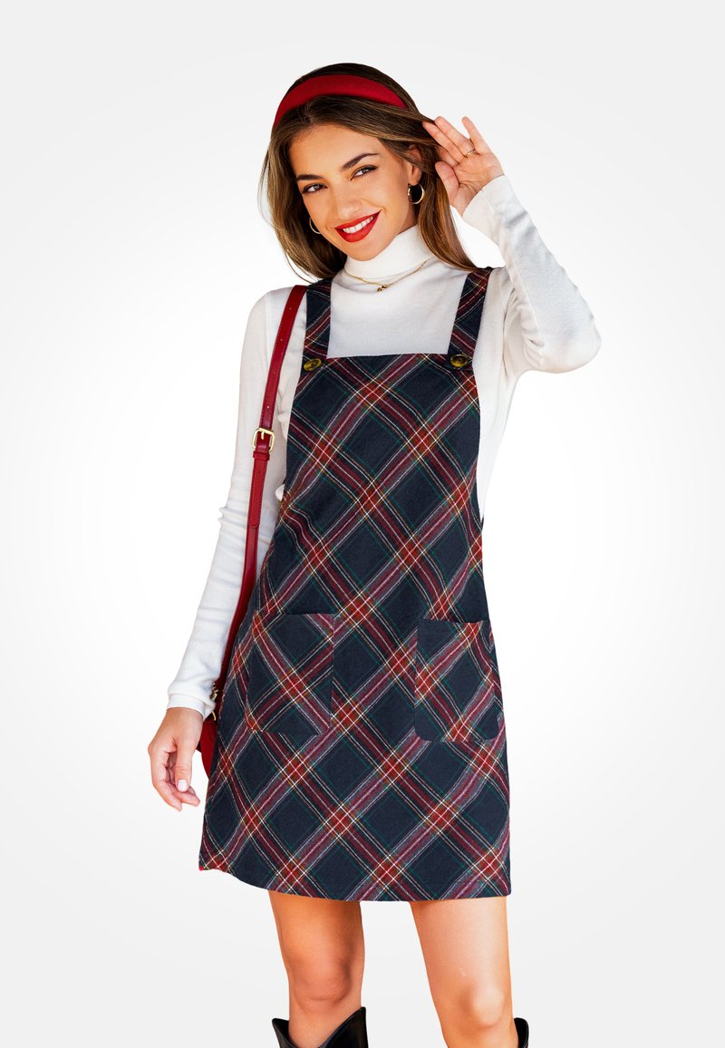 Vestido de tirantes a cuadros en tonos azul marino con acentos en rojo y verde, que cuenta con dos bolsillos delanteros y tirantes ajustables. Llevado sobre un cuello de tortuga blanco.