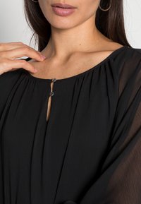 Czarna bluzka z okrągłym dekoltem, plisowanym materiałem i dwoma srebrnymi zatrzaskami na klatce piersiowej. Lekka, zwiewna struktura.