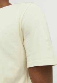 Camiseta de manga corta de color beige claro, hecha de tela suave, con un cuello redondo y costuras limpias en las mangas y el dobladillo.