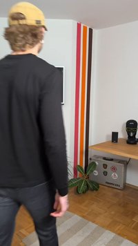 Mustat pikad varrukatega särk ja tumedad teksad, mida kannab inimene, taustal pruun puidust laud, roheline taim ja värviline triibuline sein.