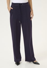 Pantalons larges violets avec une taille à cordon, des poches latérales et une texture lisse, assortis à des escarpins noirs à bout pointu.