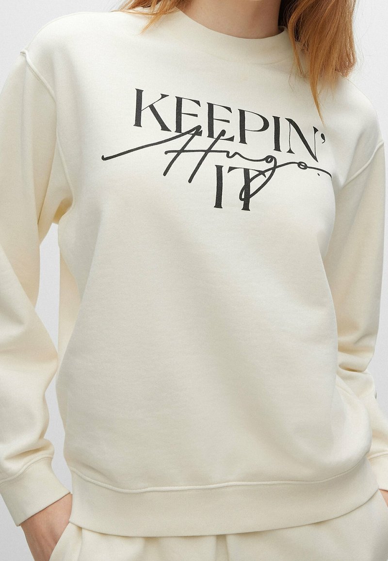 Sweat-shirt crème avec texte noir indiquant "KEEPIN' IT", porté par une personne à la peau claire et aux cheveux roux, mains dans les poches.
