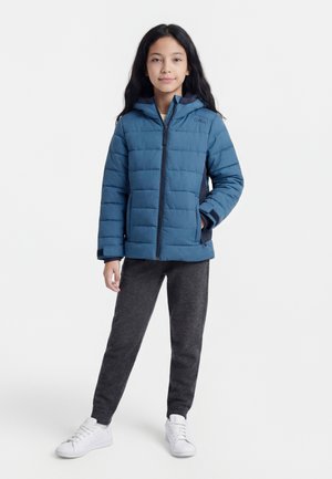 Dievča s dlhými tmavými vlasmi, ktoré má na sebe modrú prešívanú bundu, tmavosivé tepláky a biele tenisky, stojí s jednou rukou v vrecku.